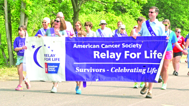 Following the Suffield High School Band, First Selectman Melissa Mack, waving, and her family lead the Survivors and Caregivers lap at the start of this year’s Relay For Life. From the left at the banner are Survivor Elizabeth Mack and her caregivers: her mother Melissa, her brother Andrew (peeking over the banner), and her father David.