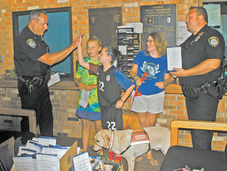 Holding one of the gift bags that Kayleigh Mahannah and Leah Beaudoin had brought to the Police Station, Officer Jeff Reynolds exchanges a high five with Leah. At the right are Amy Reay, who came with the girls, and Officer John Lacic, with another bag. That’s Amy’s service dog, Delancey, sniffing at the box of gift bags.