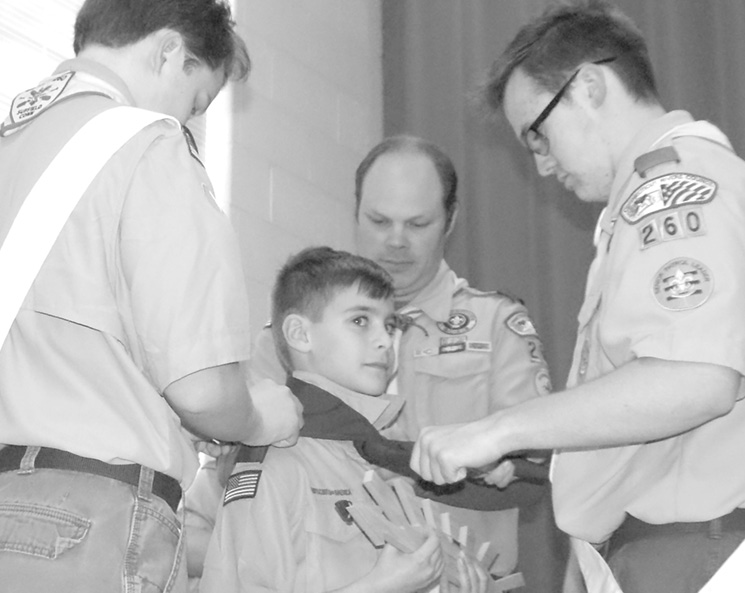 During the final step of Pack 266's Blue and Gold Celebration, Webelos Cub Scout Reid Sunshine looks up patiently as three members of Boy Scout Troop 260 outfit him with a neophyte Boy Scout neckerchief. Like ten other Cubs standing nearby, he is holding the mounted Arrow of Light symbolizing his successful completion of the full Cub Scout program. Surrounding Reid, from the left, are: Life Scout John Cremmins, Scoutmaster Mike Cremmins, and Senior Patrol Leader Mickey Barron. The ceremony, which was followed by a late afternoon dinner, took place in Father Ted Hall on March 4.