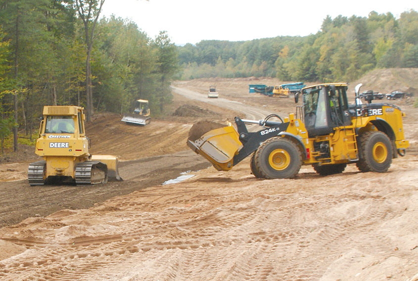 A front loader approaches with a new bucket of road material as a caterpillar bulldozer spreads the previous load on the route of the new roadway being constructed in the old sand pit. In the middle distance, another bulldozer is leveling a roadside region. The graded route of the new road can be seen in the distance.