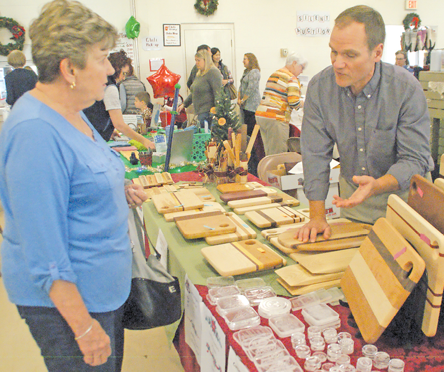 Joe Grimard explains to Joyce LaPoint of Westfield how Suffield High School’s new laser tool, bought with donations, was used to engrave and cut the wide array of useful and decorative wooden items he was selling at Sacred Heart Church’s Father Ted Hall. Sales proceeds support the Earth and Outing Club he advises at SHS.