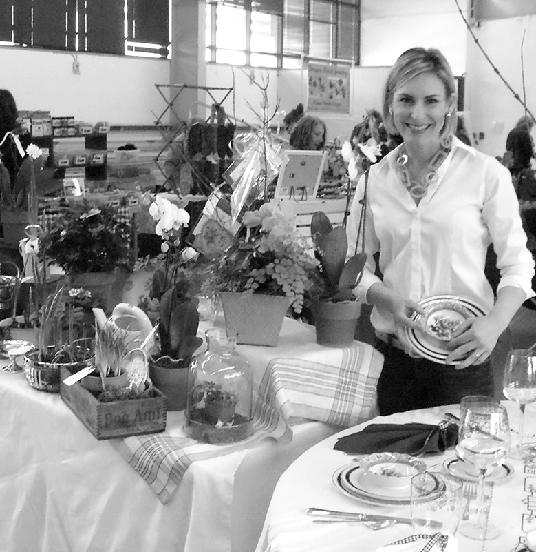 Local gardener and television personality Julie Harrison holds a sample of the complete table setting for six she offered in an unusual silent auction at the February 10 Winter Farmers’ Market. At the left is the array of potted flowers, succulents and cactuses she more commonly produces. The market on February 10 was the last of the season.